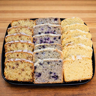 The Breakfast Loaf Platter features three rows of sliced cakes—blueberry, cinnamon swirl, and plain with icing—arranged neatly on a black tray atop a wooden surface.