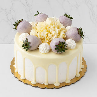 The White Chocolate Gateau features white chocolate ganache drips, light purple-dipped strawberries, French buttercream swirls, and white chocolate curls on a gold cake board, set against a marble background.