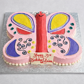 Our Butterfly Wings cake features pink, yellow, blue, and purple icing with swirls and a smiling face. A plaque reads "Happy 3rd Birthday Elaina" in red icing. Contact us for pricing.