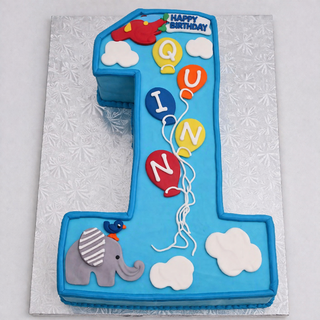 The Elephant Number cake is shaped like the number 1 and features clouds, balloons spelling QUINN, an elephant, a bird, and a red airplane with a Happy Birthday banner. This blue personalized cake showcases white and colorful decorative details.