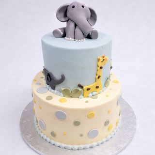 The Elephant Safari cake features two tiers with pastel polka dots, fondant giraffe, lion, and elephant decorations, topped with a large gray fondant elephant—an adorable baby shower choice. Presented on a silver board.