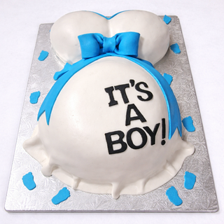 The Mamma Belly cake is shaped like a pregnant belly with a blue bow and "ITS A BOY!" written on it, decorated with blue fondant baby footprints and elegantly presented on a silver tray.