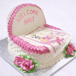 A baby shower cake shaped like a bassinet, decorated with pink and white icing, pink roses, "WELCOME BABY" on the headboard, and topped with a colorful animal-themed Bassinet Blanket.