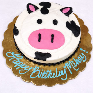 Cow Smash is a round custom cake made to look like a cow's face, with pink ears and nostrils, black spots, and "Happy Birthday Mikey!" in blue frosting on a gold board—perfect for birthdays or baby showers.