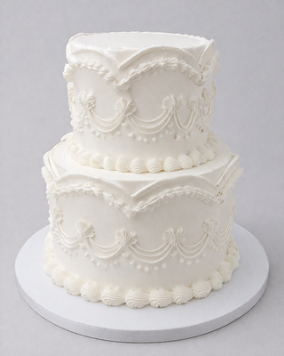 The White Bridal is a two-tiered white cake with intricate piped icing, scalloped edges, swags, and dots, displayed on a white cake board against a light gray background.