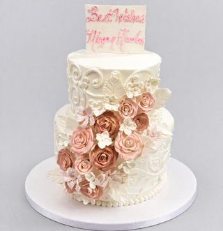 The Cascade Tier is a two-tier white cake adorned with ornate icing, blush pink and white sugar roses, leaves, and a rectangular plaque on top that reads "Best Wishes Mary & Andre" in pink icing. Custom pricing available upon request.