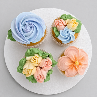 The Flower Assorted features four cupcakes on a white round plate, each topped with pastel buttercream floral designs in blue, pink, yellow, green, and orange.