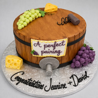 The "Wine Barrell" cake is topped with fondant grapes, cheese, and a corkscrew. A sign reads “A perfect pairing” and the board says “Congratulations Jeanine + David.” Custom flavors and sizes are available.