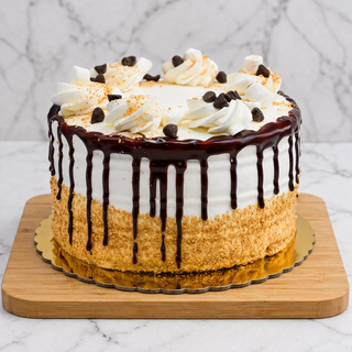 The S'mores Layer Cake is a round cake with white buttercream frosting, chocolate ganache drips, a crumb-coated base, and topped with whipped cream swirls, chocolate chips, and mini marshmallows.