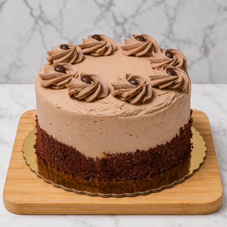 The Cappuccino Layer Cake features light brown mocha buttercream frosting, topped with swirls and chocolate pieces. It sits on a wooden board with chocolate crumbs at the base, against a marble background.