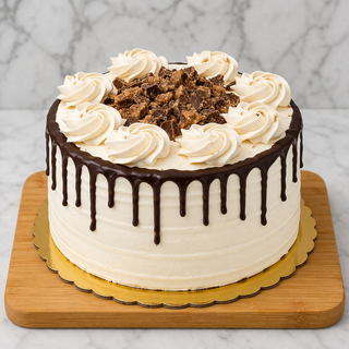 The Peanut Butter Layer Cake features a round shape, white frosting, chocolate ganache drips, swirls of frosting on top, and chopped chocolate candy pieces in the center, presented on a wooden board.