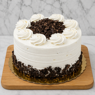 The Oreo Layer Cake features white frosting, whipped cream swirls, and chocolate crumbles on top. It rests on a gold cake board and wooden tray, displayed against a marble background.