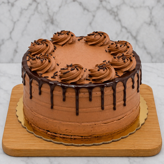 The Chocolate Layer Cake features smooth chocolate frosting and a rich ganache drip, topped with swirls of chocolate frosting and sprinkles, presented on a wooden board.
