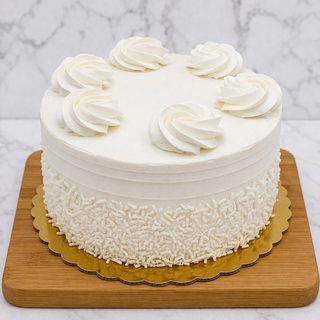 The White Layer Cake is a round cake with white buttercream, topped with six white icing swirls and white sprinkles along the bottom edge. It’s presented on a gold cake board on a wooden tray, set against a marble background.