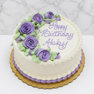 A Decorated Layer Cake: round, white, with purple icing roses, green leaves, and "Happy Birthday Abby" in purple buttercream on a gold cake board against a marble background.