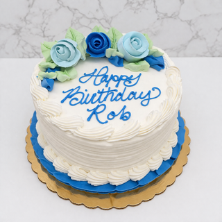 The Decorated Layer Cake features a round chocolate base with white buttercream frosting, blue and green icing roses, blue and white piped borders, and “Happy Birthday Rob” in blue icing on a gold cake board for a custom look.