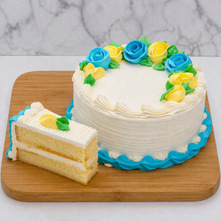 A Gluten Free Cake with two white-frosted layers, decorated with blue and yellow icing roses, sits on a wooden board. A slice is cut and placed nearby, and blue icing trims the base of this delightful treat.