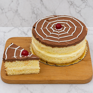 A Boston Cream Pie with chocolate icing, a white spiderweb pattern, and a cherry on top rests on a wooden board beside a sliced piece showing its creamy filling.
