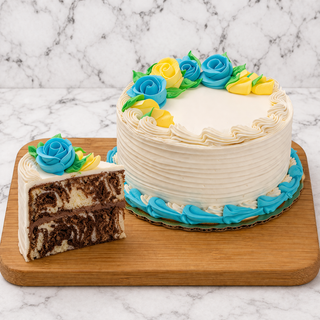 The Marble Swirl Cake, topped with buttercream and blue, yellow, and green icing flowers, sits on a wooden board. A single slice rests beside it, showing the marbled interior.