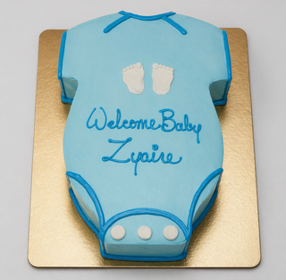 The Onesie Cutout cake is shaped like a blue onesie with white feet prints and "Welcome Baby Zyaire" in blue icing, perfect for baby showers. It's displayed on a gold board.
