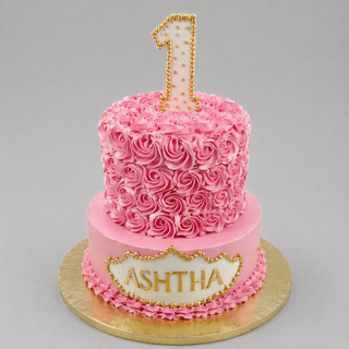 The Rossette Number is a two-tier pink birthday cake with rosette number icing, a gold-beaded number one topper, and "ASHTHA" on a plaque. Ideal for baby showers, it sits elegantly on a gold board.