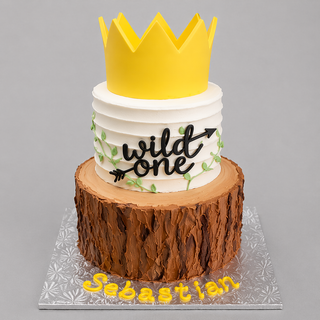 The Wild One cake features two tiers: a tree trunk base, white top with green vines, yellow crown topper, “wild one” in black, and “Sebastian” in yellow on the board—an ideal birthday centerpiece.