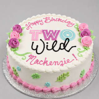 Two Wild round white cake decorated with pink, purple, and green frosting flowers, topped with the message “Happy Birthday TWO Wild Mackenzie!” in colorful icing.