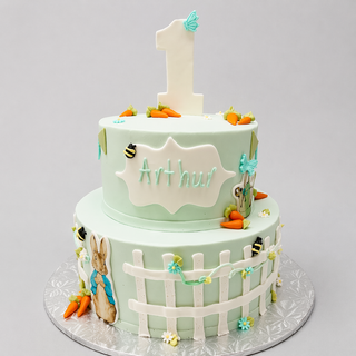The Peter Rabbit Tier is a two-tier pastel green cake with carrots, bees, a picket fence, and a bunny, “Arthur” on the side, and a large white number 1 topper. Presented on a silver board—perfect for birthdays.