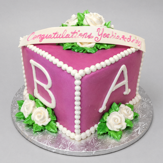 A Single Block cake in a hexagonal shape with purple icing, white icing roses, green leaves, large white "B" and "A" on the sides, and a ribbon on top that says "Congratulations Yoshi & Lindy.