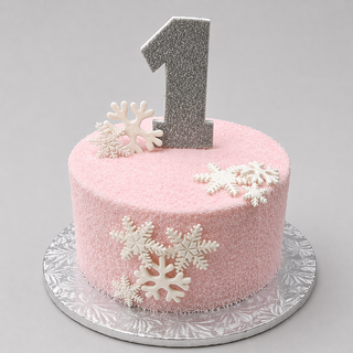 The Winter One cake is a pink custom birthday cake with white snowflakes and a large silver glitter number 1 topper, presented on a silver patterned cake board.