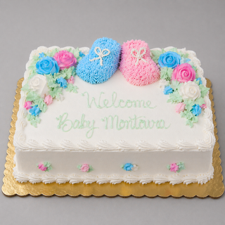 A rectangular white baby shower cake topped with colored booties, pink and blue roses, green leaves, and “Welcome Baby Montava” in green icing, presented on a gold board. Product: Colored Booties.