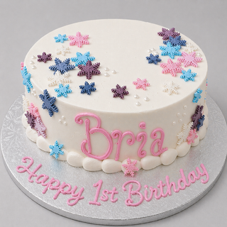 The Snowflake Cascade cake is a round birthday cake with white pearls, a snowflake cascade, "Bria" written in pink icing on the side, and "Happy 1st Birthday" in pink on a silver cake board.