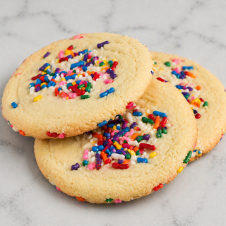 Three Large Sprinkle Cookies, inspired by the classic American recipe, are topped with vibrant rainbow sprinkles and stacked on a light marble surface.