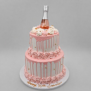 Champagne Tier: Two-tier white cake with pink drip icing, pink and white swirled frosting, gold accents, a mini rosé champagne bottle topper, and “Happy 70th Joanne” in gold on the cake board.