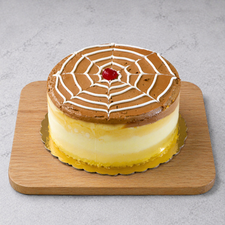 The GF Boston Cream Pie is a round vanilla yellow cake with a caramel top, white spiderweb pattern, and a red cherry in the center, featuring gluten-free yellow and white layers and served on a wooden board.