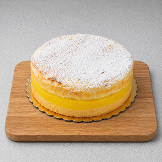 The GF Lemon Delight Cake, a round gluten-free lemon cake with tangy lemon preserves, thick yellow filling, and powdered sugar dusting, is displayed on a wooden board against a light gray background.