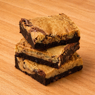 Three stacked GF & Dairy Free Brookies—a dessert with a dark chocolate brownie base and golden cookie-like top—rest on a wooden surface. This treat combines chewy brownie and chocolate chip cookie layers.