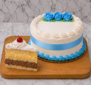 A round Rum Cake with blue roses, a blue ribbon, and white frosting rests on a wooden board. A front slice displays chocolate, vanilla, and yellow layers filled with Italian custard, whipped cream frosting, and topped with a cherry.