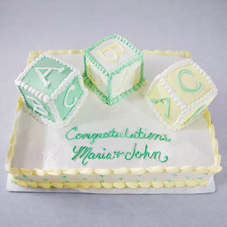 The "Baby Blocks" cake is a rectangular treat decorated with pastel green and yellow baby blocks, finished with white icing and the message “Congratulations Maria & John” in green—perfect for a baby shower.