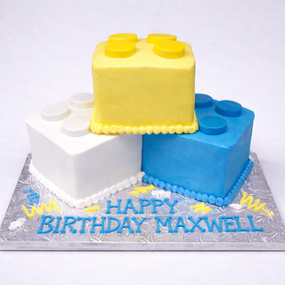 Three custom Lego Blocks cakes in yellow, white, and blue are stacked on a silver board with "Happy Birthday Maxwell" written in blue and yellow frosting—a fun treat for kids.