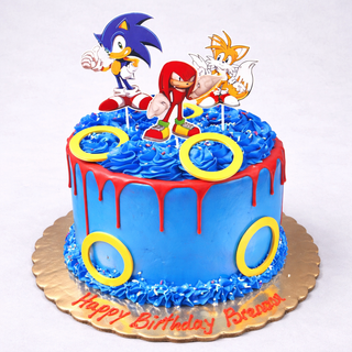 A round blue cake with red icing drip and yellow rings, topped with Sonic Toppers and characters like Knuckles and Tails. The cake board reads “Happy Birthday Praneesh” in red icing—perfect for kids!.