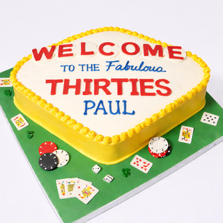 The Vegas Sign cake is a square, sports & hobby-inspired cake with playing cards, poker chips, and a Vegas motif. “Welcome to the Fabulous Thirties Paul” decorates the top. It rests on a green base. Custom pricing available.