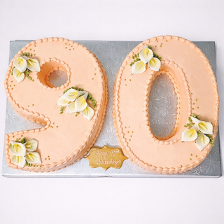 Celebrate with our Callalily Number cake: a peach-hued, number 90 design adorned with white and yellow flowers on a silver board, featuring a "Happy 90th Birthday" plaque. Contact us for pricing.