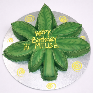 The Marijuana Cutout cake is shaped and decorated like a green marijuana leaf with "Happy Birthday Hi MiLLSz" in yellow icing, yellow swirl accents, and presented on a round silver board. Custom pricing available for this unique design.