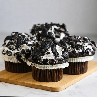 Four Oreo Creamy Dreamy Cupcakes, topped with buttercream frosting and crumbled Oreo cookies, are arranged on a wooden board against a light gray background.