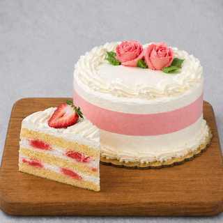 The GF Strawberry Shortcake, a round vanilla gluten-free cake with white and pink frosting, is topped with two pink roses and green leaves. On a wooden board, a slice shows layers of sponge, homemade whipped cream, and strawberries.
