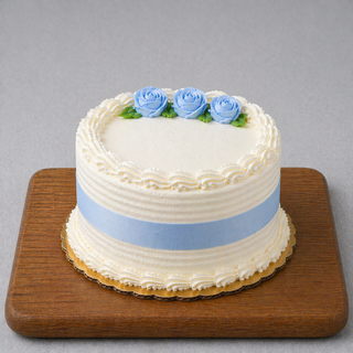 The Traditional Whip Cream Cake is a round homemade cake with white whip cream frosting, adorned with a light blue ribbon and three blue buttercream roses with green leaves—ideal for adding your favorite specialty cake fillings.