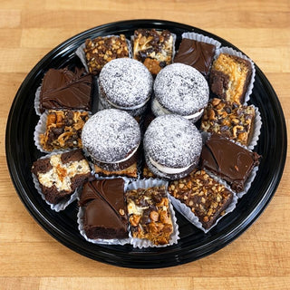 The Brownie Tray features assorted chocolate desserts like whoopie pies, mini brownies with chocolate frosting, and nut-topped bars, all arranged neatly on a black plate set atop a wooden surface.