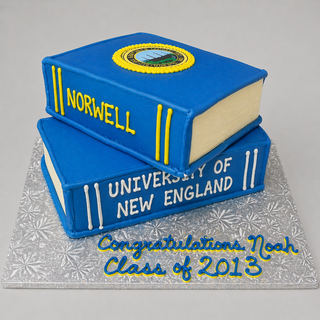 A Double Book graduation cake labeled Norwell and University of New England sits on a silver board with "Congratulations Noah, Class of 2013" in blue icing.