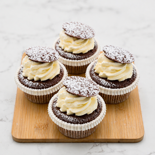 Four Chocolate Drop Cupcakes with whipped cream swirls, topped with round chocolate discs and powdered sugar, arranged on a light wooden board against a white background.
